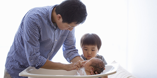 A young father of Asian descent bathes an infant in a basin while his toddler-age son looks on.
