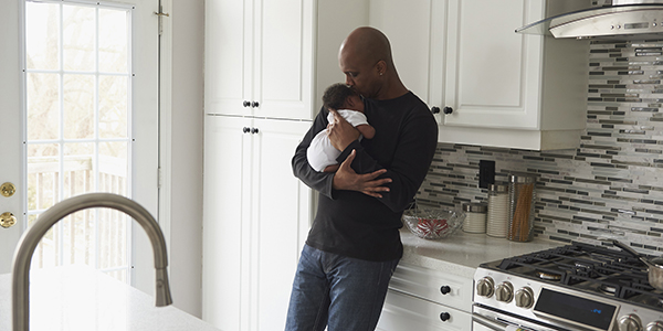 A father stands in the kitchen holding and kissing his newborn child.